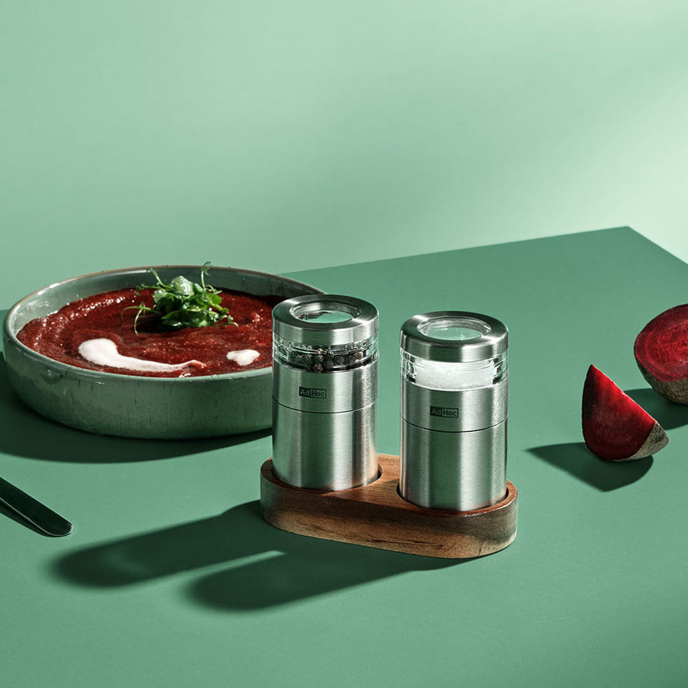 Two metal salt and pepper shakers on a wooden stand with a bowl of red soup and a beetroot on a green background.